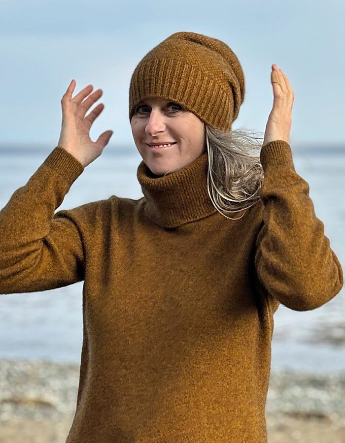 A woman wearing a warm brown ribbed knit beanie and matching brown turtleneck sweater, posing with her hands raised against a blurred beach and sea background.