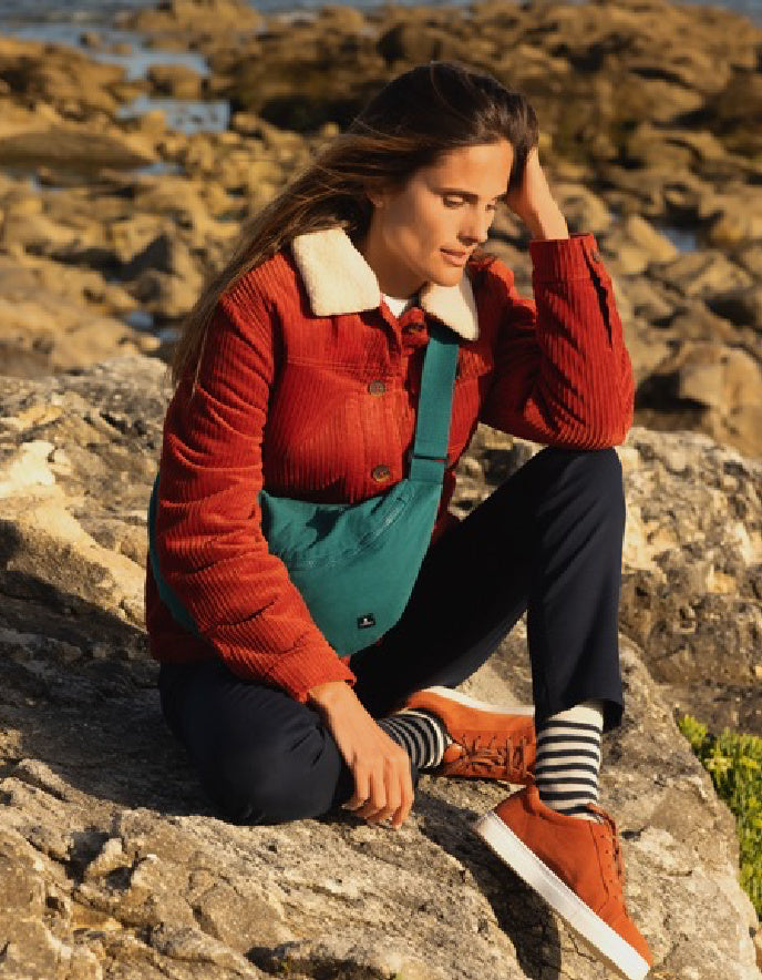 Woman sitting on rocks by the sea wearing a red jacket and blue bag.