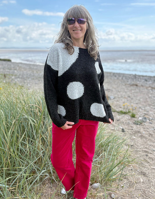 Woman wearing a black sweater with white polka dots and pink velvet pants standing on a beach.