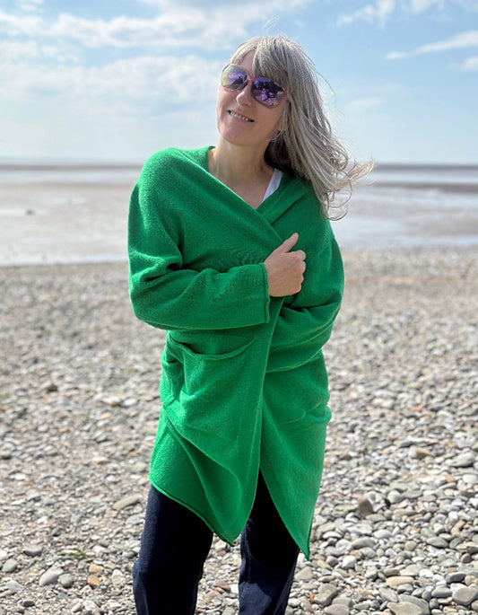 Person wearing a green wrap long merino wool cardigan on a pebbly beach with ocean and sky in the background