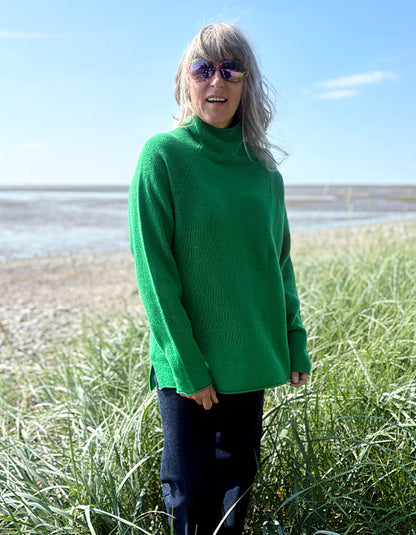 Woman wearing a green wool fisherman style sweater and sunglasses standing on a grassy field with a beach in the background.