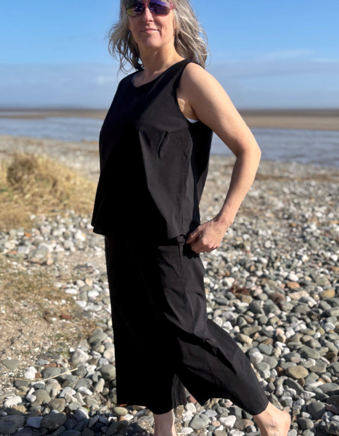 Woman in a black sleeveless dress standing on a pebbly beach with a clear sky.