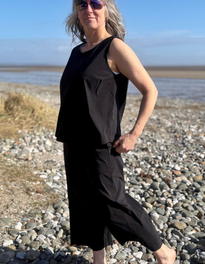Woman in a black sleeveless dress standing on a pebbly beach with a clear sky.