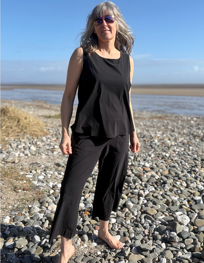 Woman in black sleeveless top and pants standing on a pebbly beach with blue sky.