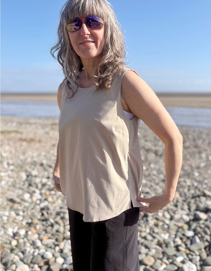 Person standing on a pebbly beach wearing sunglasses and a beige sleeveless top.