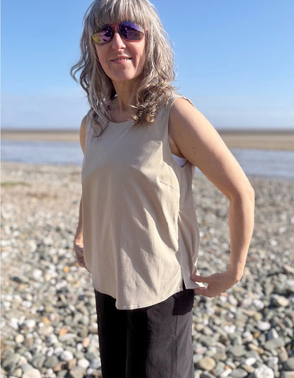 Person standing on a pebbly beach wearing sunglasses and a beige sleeveless top.