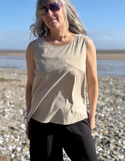Woman standing on a pebbly beach wearing a beige sleeveless top and sunglasses.