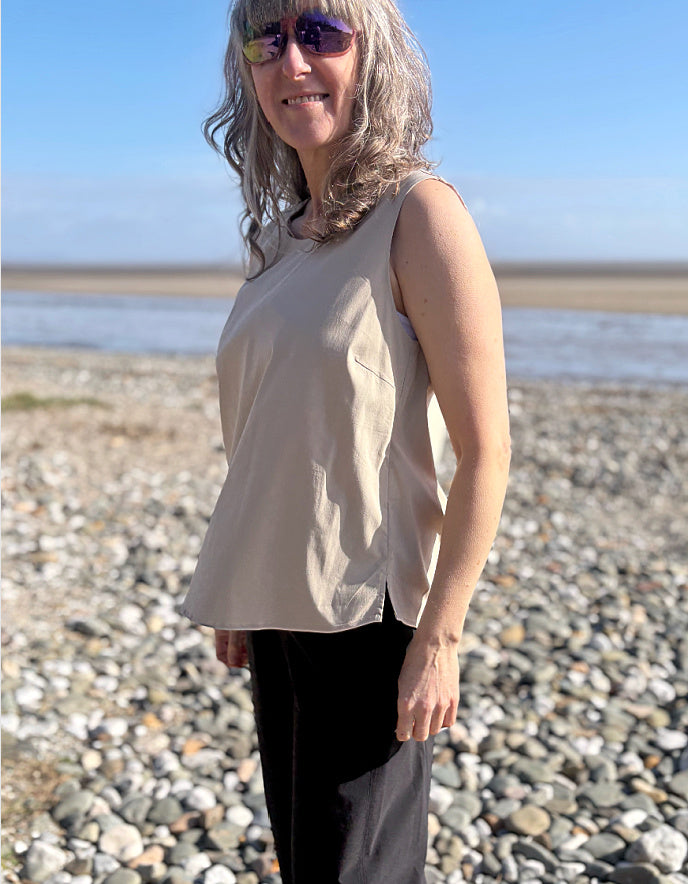 Woman standing on a pebbly beach wearing a beige sleeveless top and sunglasses.