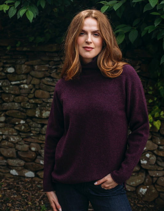Woman wearing burgundy wool turtle neck sweater standing in front of a stone wall and greenery