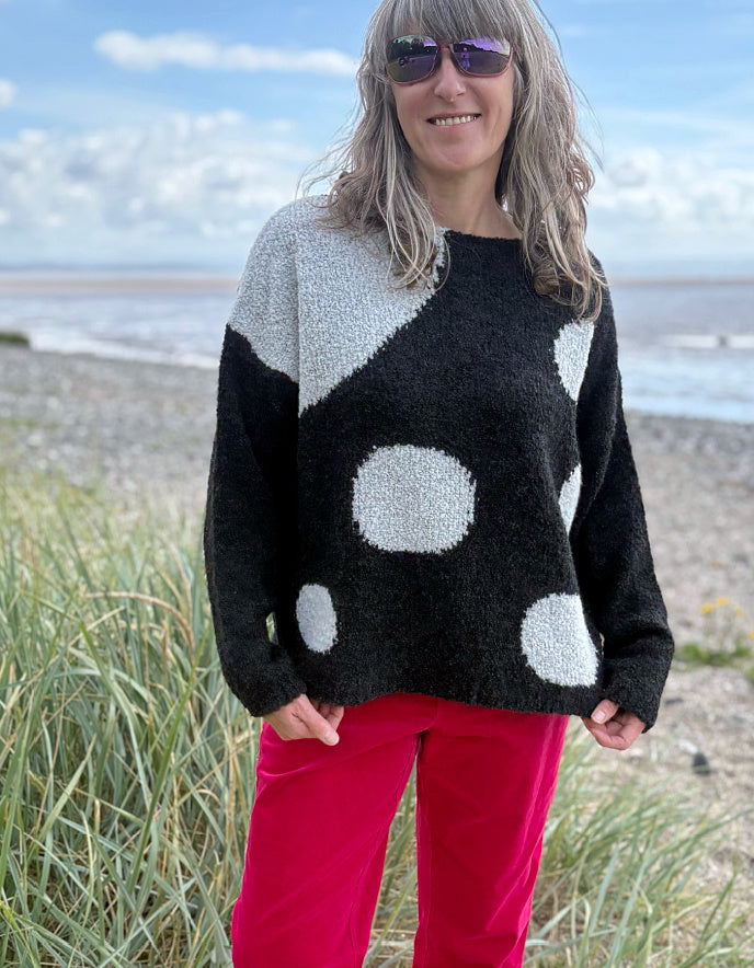 Woman wearing a black sweater with white polka dots and pink velvet pants standing on a beach.