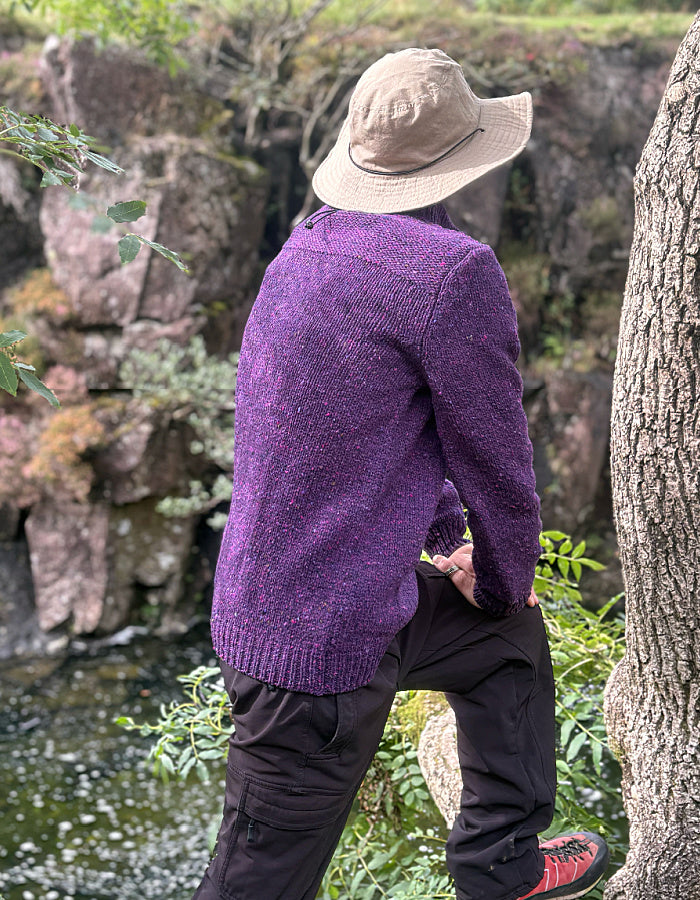 A person wearing a textured purple knit sweater, black pants, a beige bucket hat, and red shoes stands outdoors near a tree and water, viewed from behind.