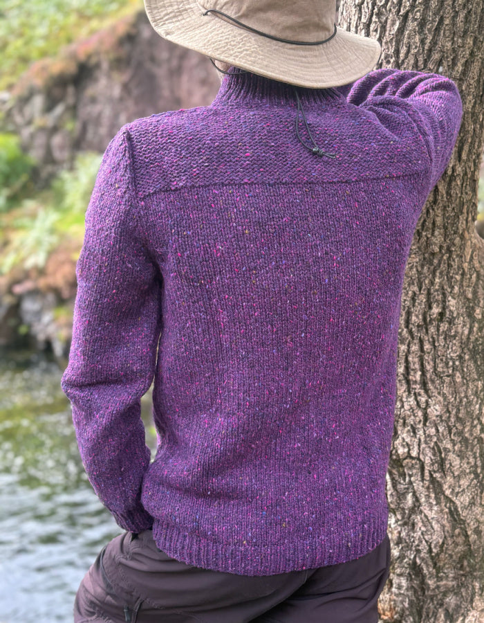 A person wearing a purple knit turtleneck sweater with flecks of pink and blue, leaning against a tree outdoors near water. The person is also wearing a beige wide-brim hat and dark pants.