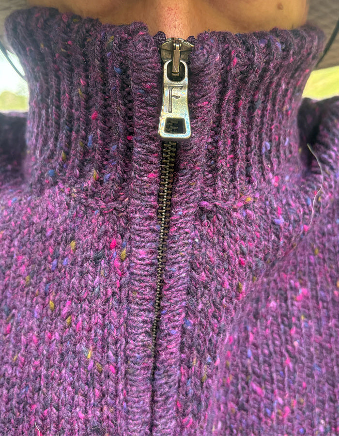 Close-up of a ribbed knit turtleneck sweater with a front zipper, featuring a heathered purple yarn with pink, blue, and yellow flecks. The silver metal zipper pull is shown.