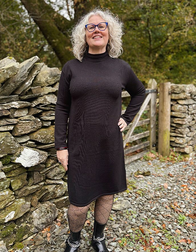 Woman wearing a black knit dress standing in front of a stone wall and wooden gate.
