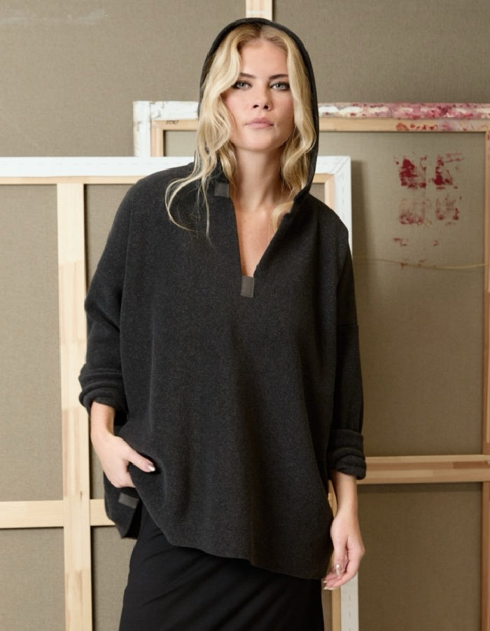 A woman wearing a dark charcoal gray hooded pullover sweater with a half-placket and side pockets, standing in front of a rustic wooden and canvas backdrop.