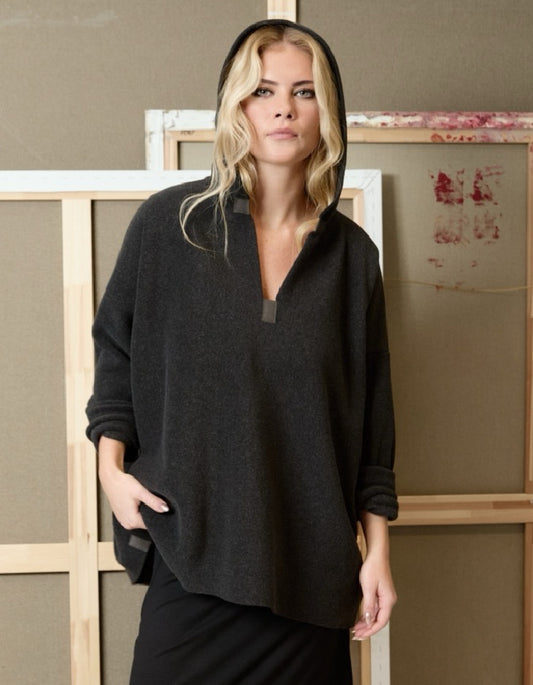 A woman wearing a dark charcoal gray hooded pullover sweater with a half-placket and side pockets, standing in front of a rustic wooden and canvas backdrop.