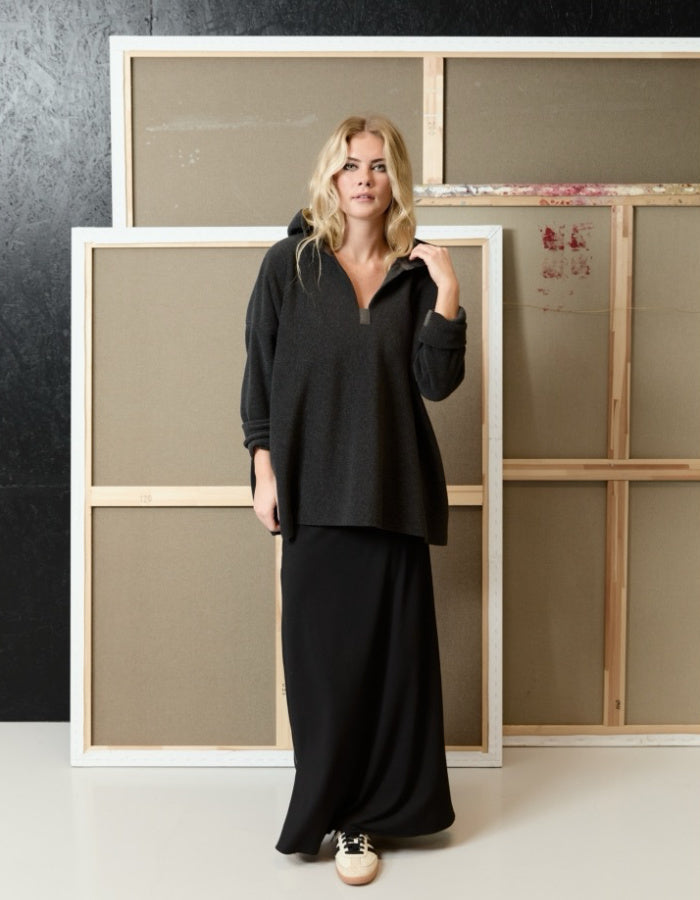 A woman stands in a studio wearing an oversized black long-sleeve top and a long black skirt, with two large framed canvases behind her against a dark textured wall.
