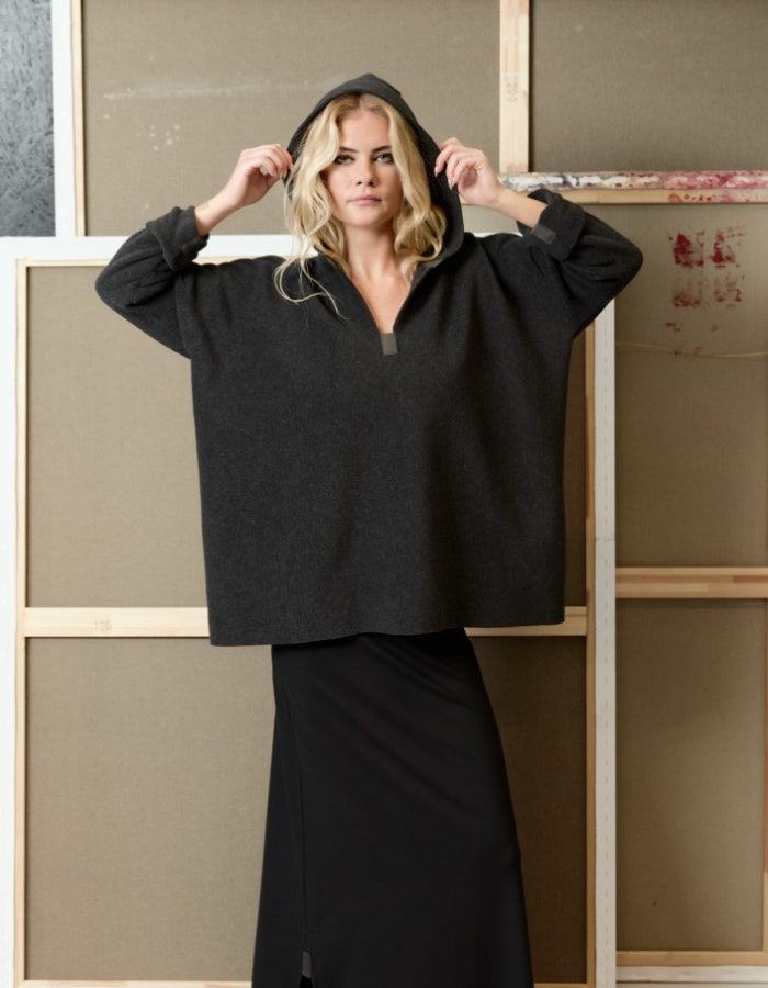 A woman wearing a dark charcoal gray cropped hoodie with a half-zip neckline and a long black skirt, posing in front of a rustic wooden and canvas backdrop.