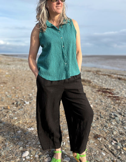 Woman standing on a beach wearing a green sleeveless top and black pants.