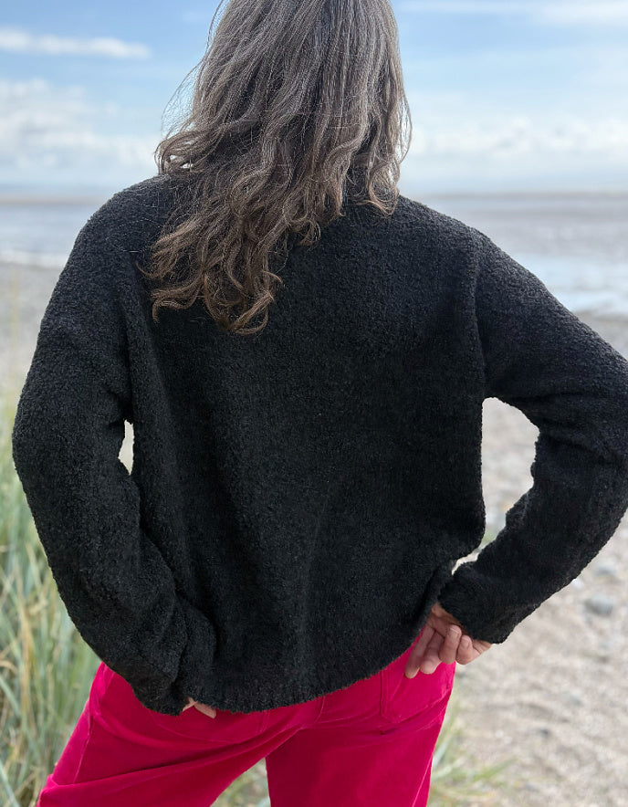 Woman wearing a black sweater with white polka dots and pink velvet pants standing on a beach.