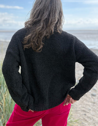 Woman wearing a black sweater with white polka dots and pink velvet pants standing on a beach.