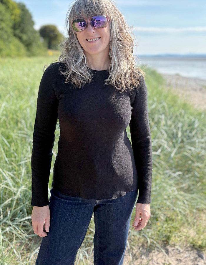 Person wearing a black long-sleeve shirt and sunglasses standing outdoors with grass and water in the background