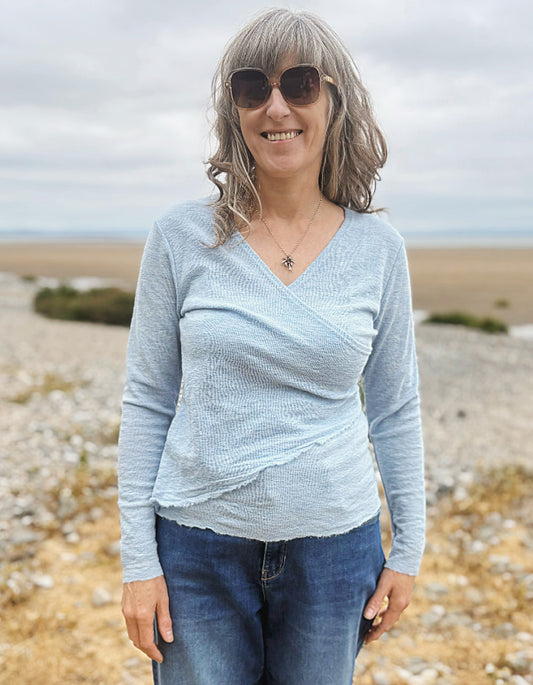 A woman with gray hair and sunglasses wearing a light blue textured long-sleeve wrap top and blue jeans, standing outdoors on a rocky beach path.