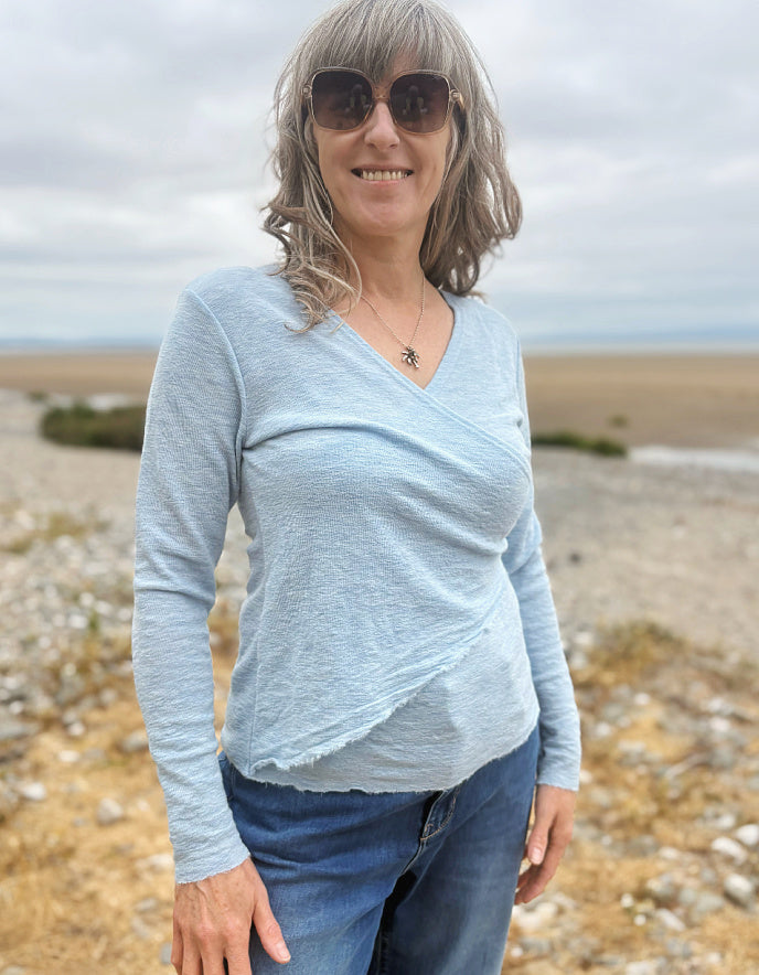 A woman with gray hair and sunglasses wearing a light blue long-sleeve wrap-style top and blue jeans, standing outdoors on a rocky beach or shoreline under an overcast sky.