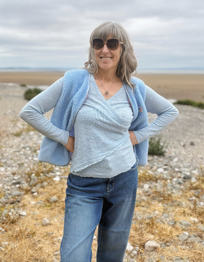 A woman wearing a light blue textured cardigan over a matching light blue wrap top and blue jeans, standing outdoors on a rocky, dry landscape under an overcast sky.