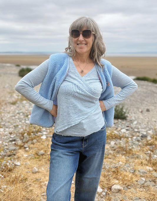 A woman wearing a light blue textured cardigan over a matching light blue wrap top and blue jeans, standing outdoors on a rocky, dry landscape under an overcast sky.