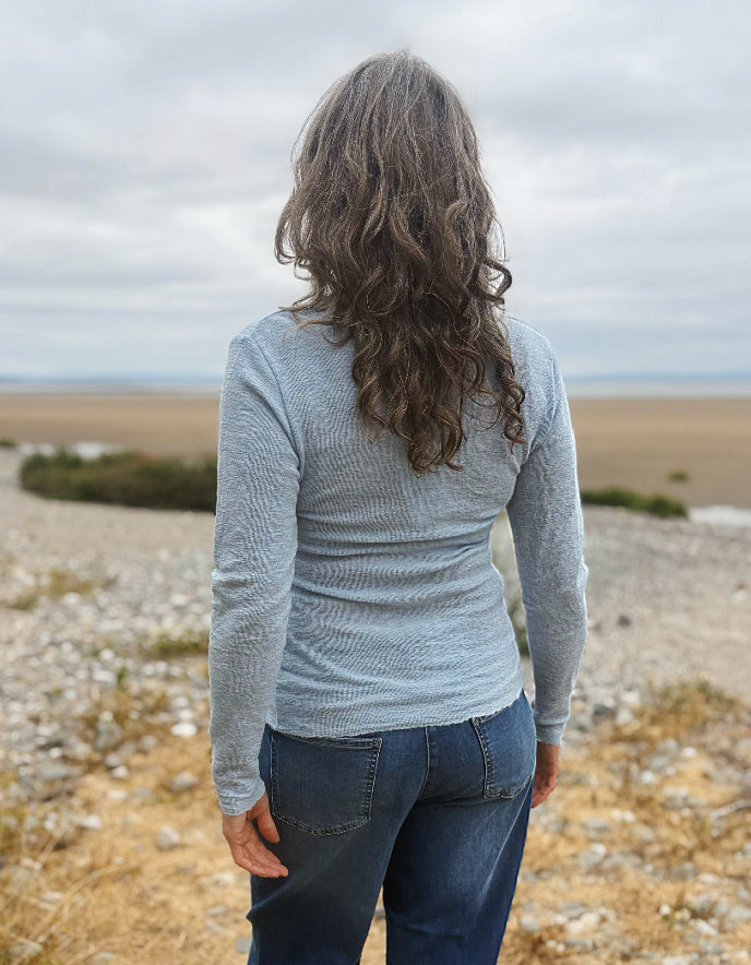 A woman with long, wavy brown hair is shown from behind, wearing a light gray ribbed long-sleeve top and dark blue jeans, standing on a rocky beach under an overcast sky.