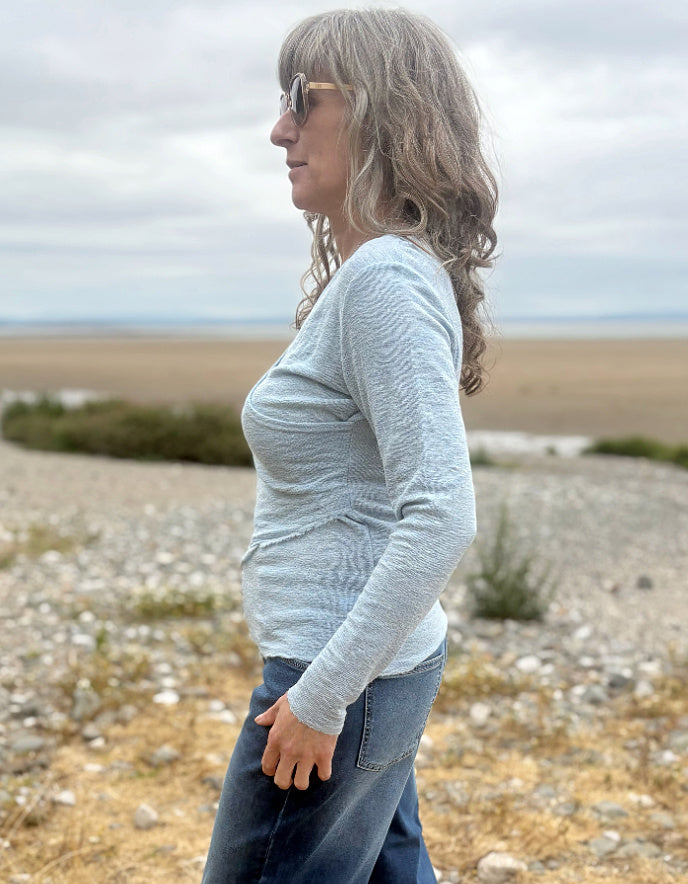 A woman with gray hair and sunglasses stands in profile outdoors on a rocky beach or shoreline, wearing a light heather gray long-sleeve top and blue jeans.