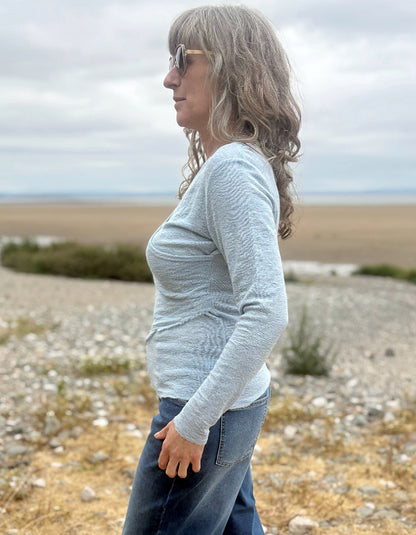 A woman with gray hair and sunglasses stands in profile outdoors on a rocky beach or shoreline, wearing a light heather gray long-sleeve top and blue jeans.