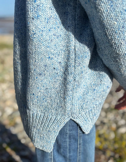 Close-up of a person wearing a light blue speckled knit sweater and blue jeans outdoors on a rocky beach.