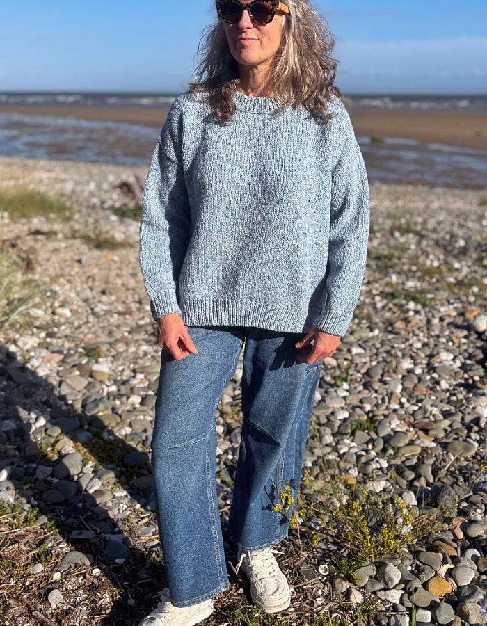 A woman wearing a light blue marled knit sweater, blue jeans, and white sneakers stands on a rocky beach with the ocean and sky in the background.