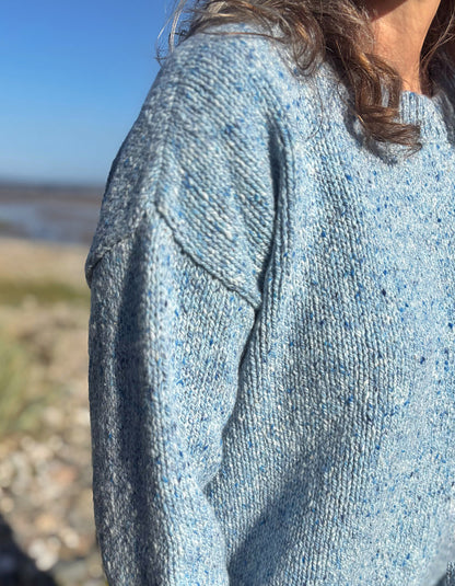 Close-up of a person wearing a light blue heathered knit sweater outdoors near a beach, with the person's shoulder, hair, and part of the neck visible.