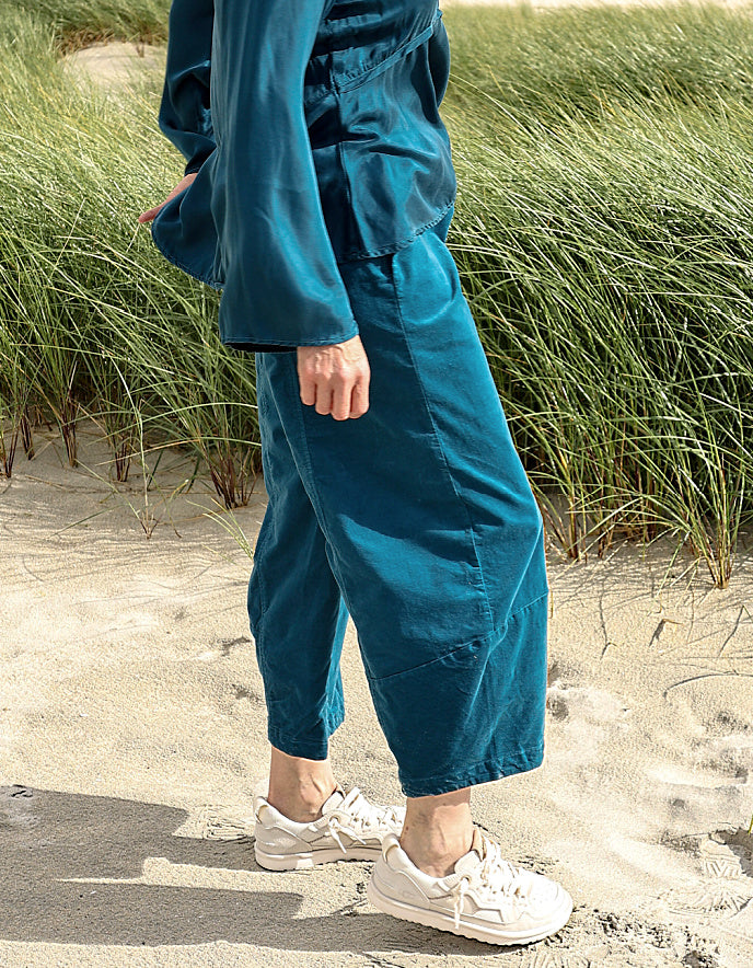A person standing on sandy beach dunes wearing loose teal-blue cropped pants and a matching top, with white sneakers, in front of tall green beach grass.