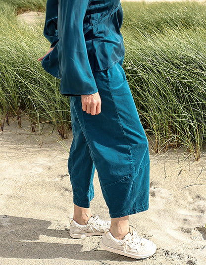 A person standing on sandy beach dunes wearing loose teal-blue cropped pants and a matching top, with white sneakers, in front of tall green beach grass.