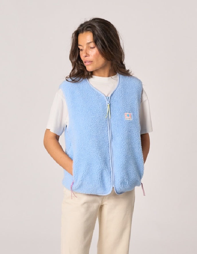 A woman wearing a light blue sherpa fleece zip-up vest over a white t-shirt and beige pants, standing against a solid light gray background.