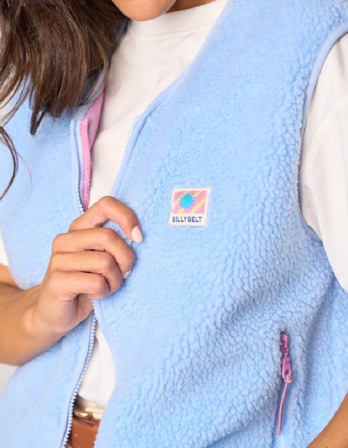A close-up of a person wearing a light blue sherpa fleece vest over a white shirt. The vest features a small square 'BILLY BELT' logo patch on the left chest and a pink zipper pull.