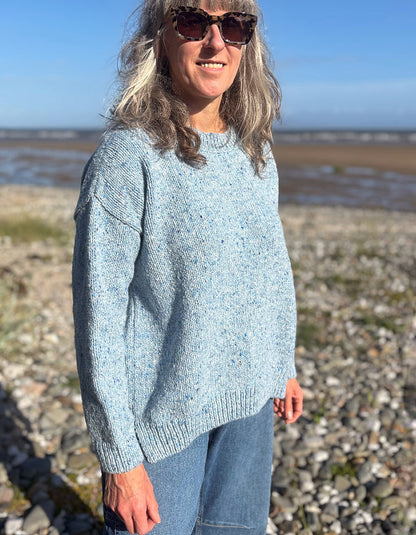 A woman wearing a light blue donegal tweed merino knit sweater and blue jeans stands on a rocky beach with water and sky in the background.