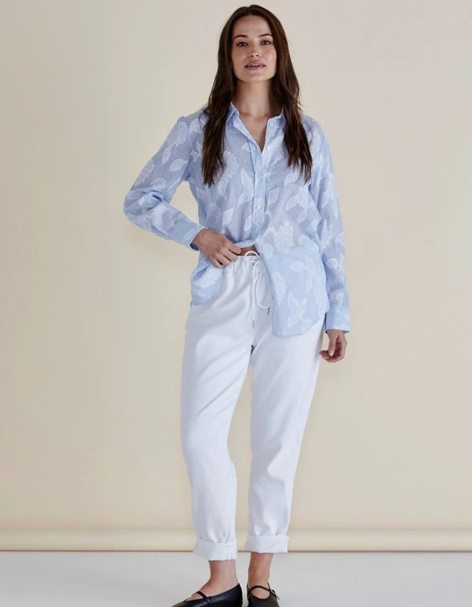 Woman wearing a light blue and white striped patterned shirt with lace floral print and white pants against a beige background
