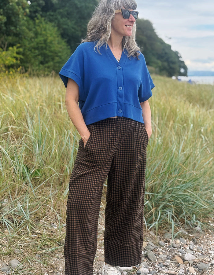 A woman standing outdoors in a grassy field near the water, wearing a bright blue short-sleeve button-front top and brown gingham wide-leg pants.