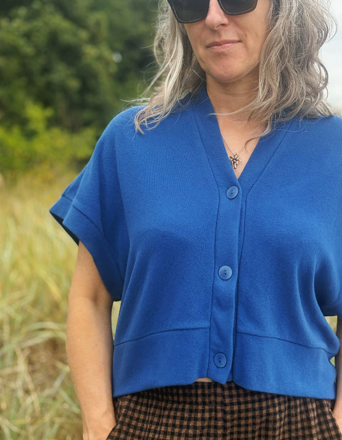 A woman wearing a bright blue short-sleeve button-front cardigan top, photographed outdoors with blurred greenery and grass in the background.
