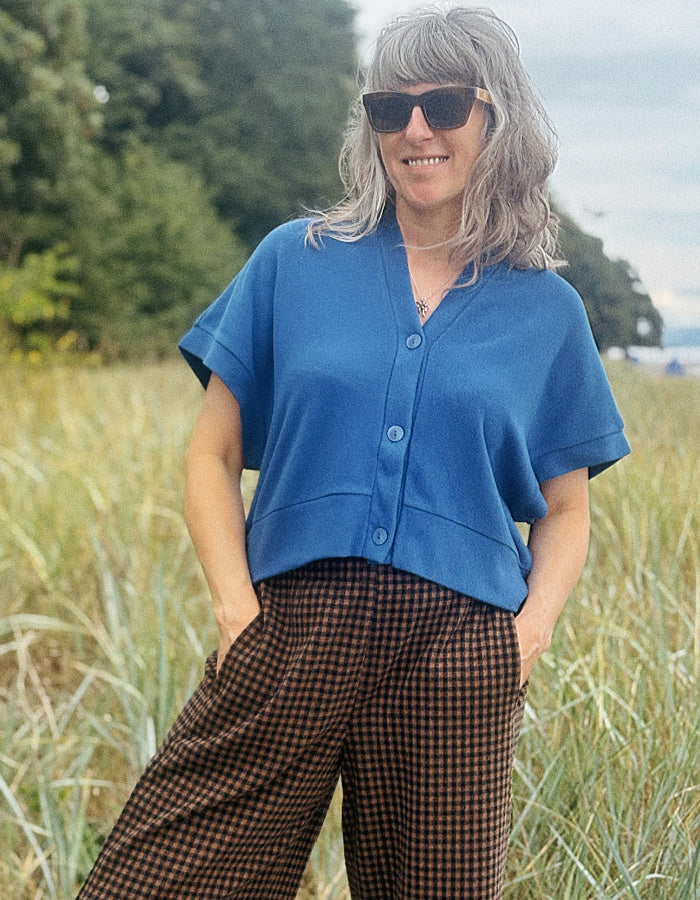 A woman with gray hair and sunglasses stands in a grassy outdoor field, wearing a short-sleeve blue button-front top and brown gingham pants.