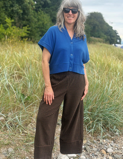 A woman wearing a blue short-sleeve button-front top and brown gingham wide-leg pants, standing outdoors in a grassy field.
