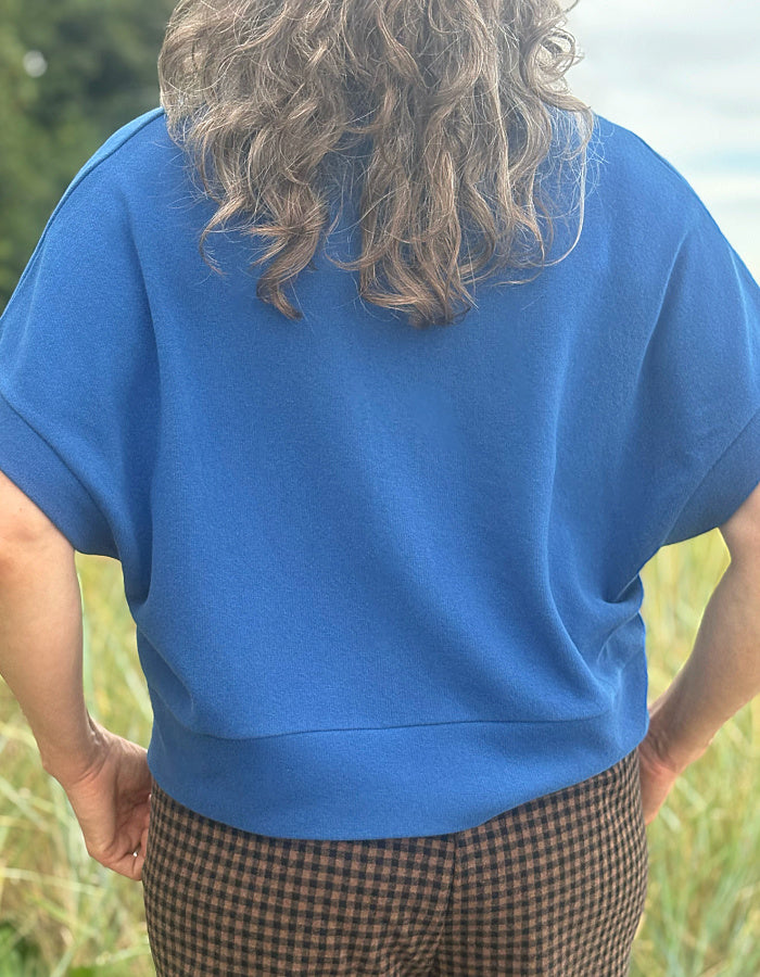 A person viewed from behind wearing a bright blue short-sleeve top and brown-and-black checkered pants, standing outdoors in a grassy field.