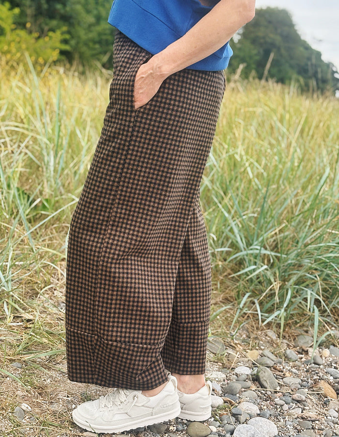 A person standing outdoors in a grassy field wearing brown and black checkered wide-leg pants, a blue top, and white sneakers.