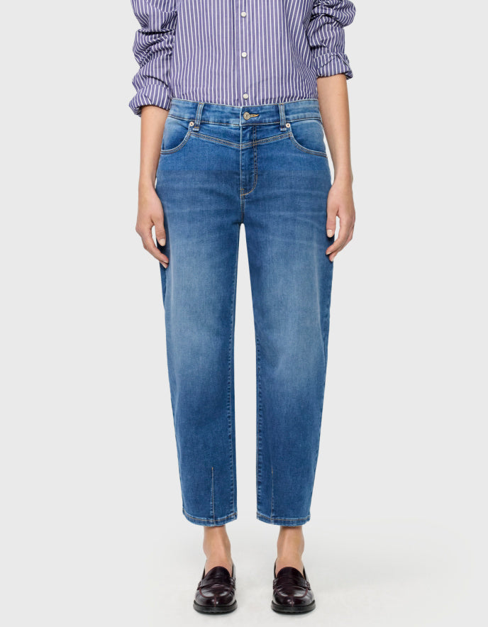 A model wearing a pair of medium-wash blue barrel jeans with a subtle vertical fade, paired with a purple and white striped shirt and dark brown loafers, against a light grey background.