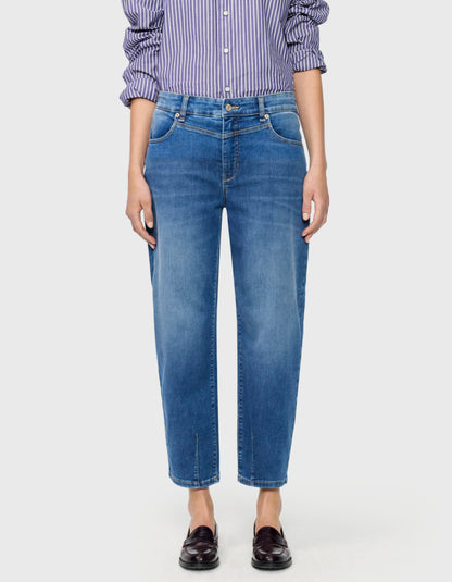 A model wearing a pair of medium-wash blue barrel jeans with a subtle vertical fade, paired with a purple and white striped shirt and dark brown loafers, against a light grey background.